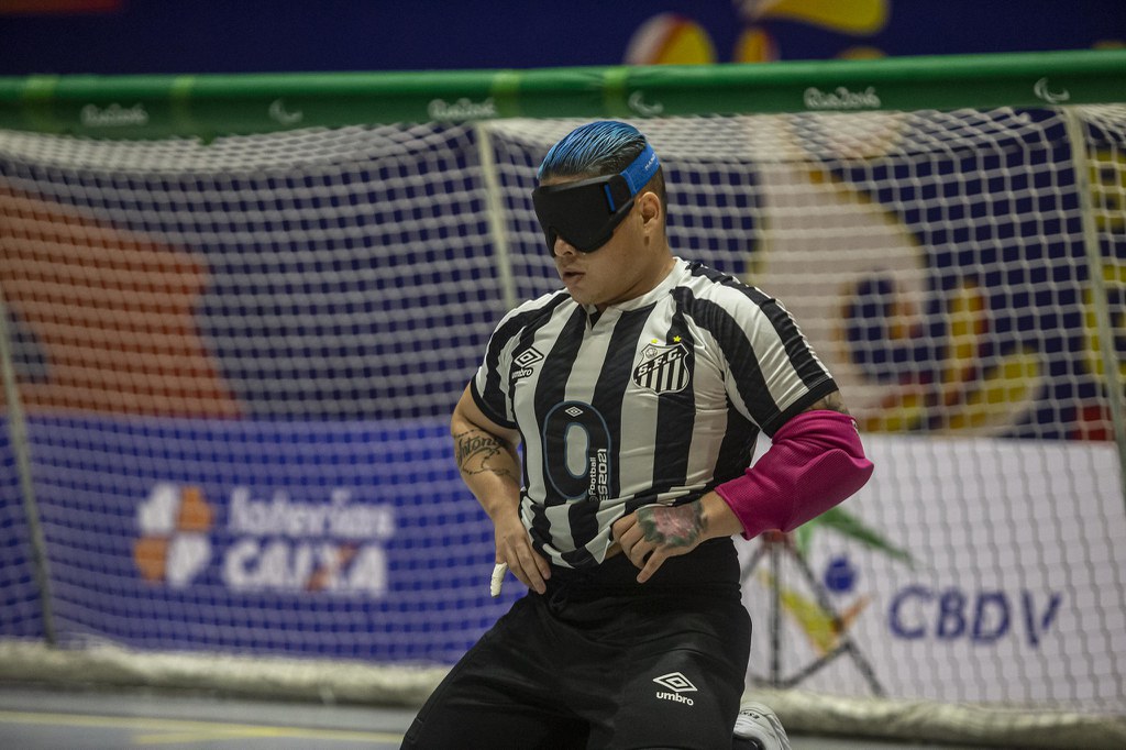 Pedrinho, do Santos, ajoelhado em posição de defesa. Ele usa uniforme listrado em preto e branco, com braçadeira rosa e tem o cabelo azul. Pedrinho, do Santos, ajoelhado em posição de defesa. Ele usa uniforme listrado em preto e branco, com braçadeira rosa e tem o cabelo azul.