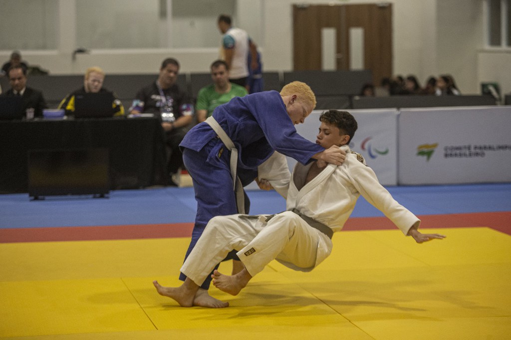 Davi Rios, de kimono azul e faixa branca, projeta Gabriel Marques (kimono branco e faixa cinza), que está caindo no tatame, com a mão espalmada. Ao fundo, há arbitragem sentada e placas do evento.