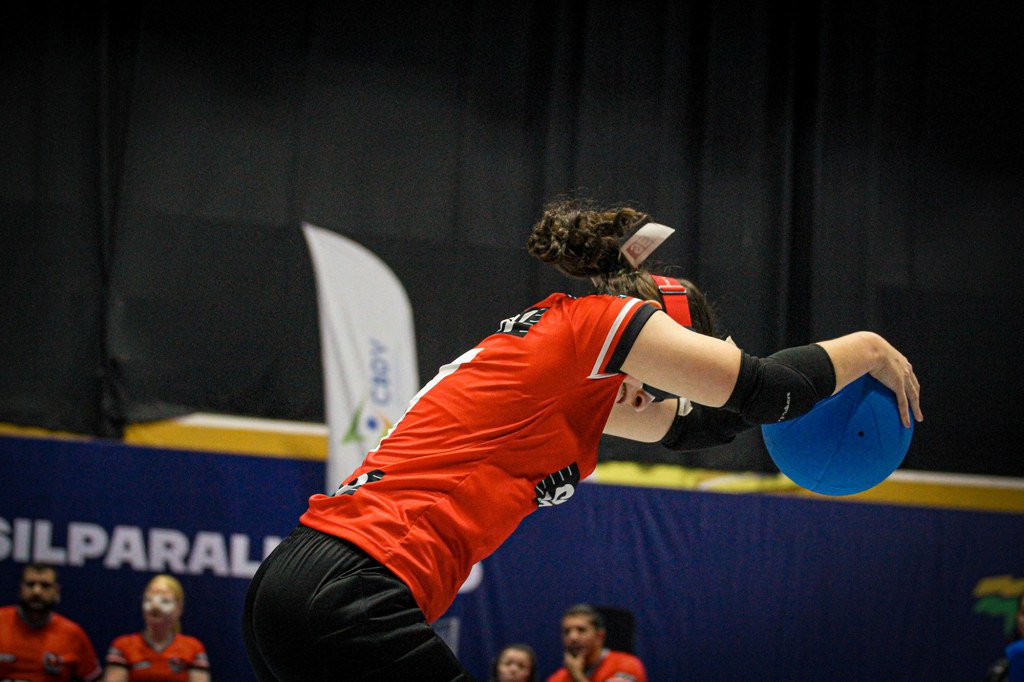 Atleta de goalball, vestindo uniforme vermelho e preto do SESI, realiza o movimento de arremesso da bola azul durante uma partida. A jogadora está inclinada para frente, com um dos braços estendido e utilizando vendas nos olhos, equipamento obrigatório da modalidade. Ao fundo, é possível ver integrantes das equipes e a estrutura da quadra de competição. Crédito: Larissa Rodrigues/CBDV.