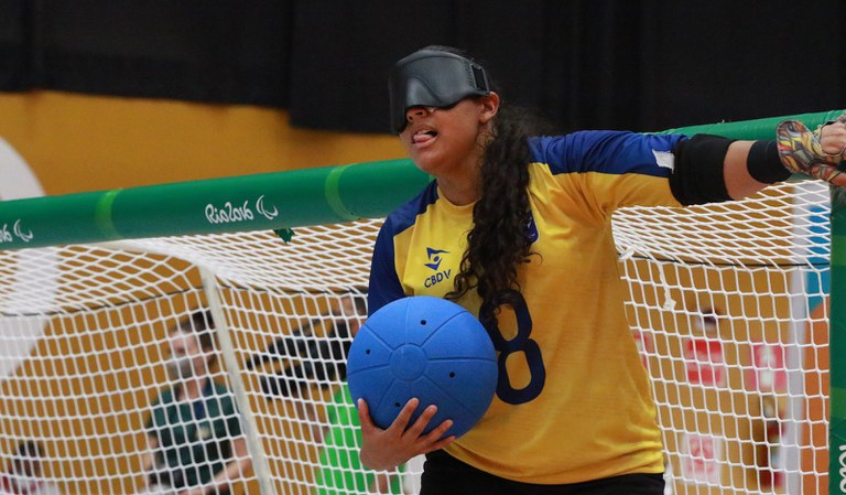 Jéssica usa a mão esquerda para tocar a baliza e se posicionar antes do arremesso. Ela é morena, tem cabelo comprido e cacheado e está com a língua para fora no momento da foto. A atleta segura a bola com a mão direita. 177A8566.JPG