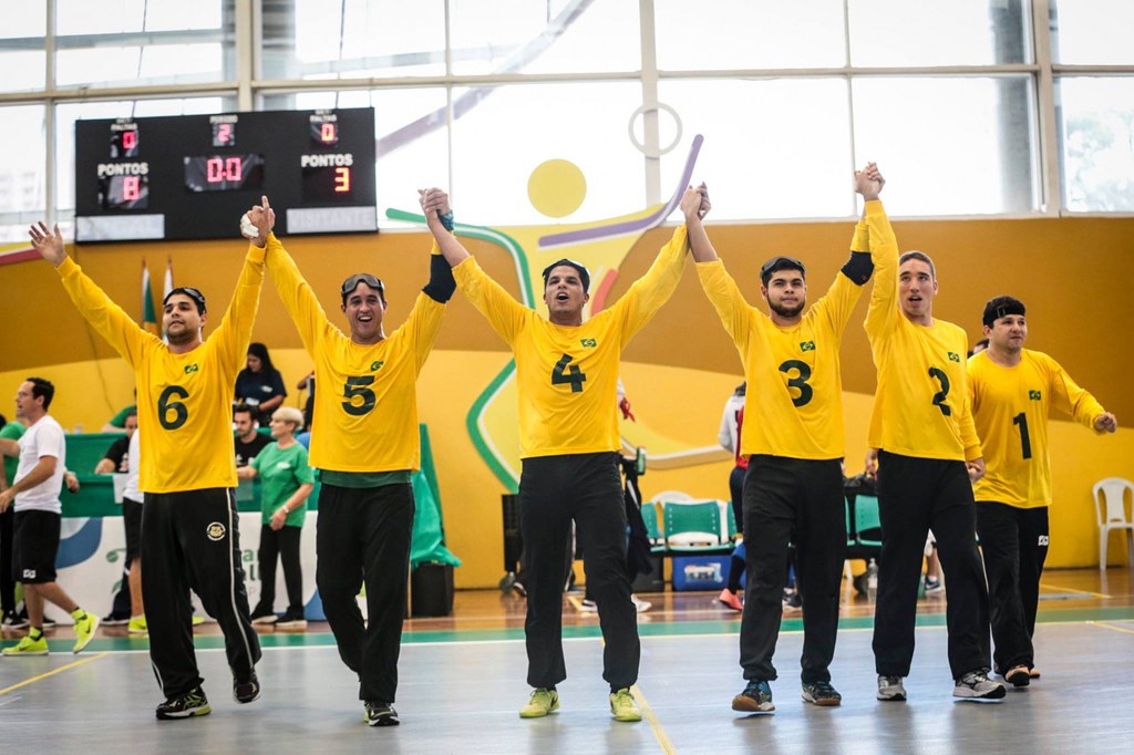 #Acessibilidade: Seleção masculina dá as mãos na quadra e ergue os braços para saudar o público durante torneio de 2017. Da esquerda para a direita, aparecem Romário, Parazinho, Leomon, Dantas, Alex e Zé Roberto, todos de camisa de manga longa amarela e calça preta.