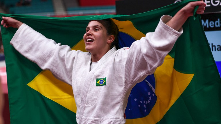 #Acessibilidade: Alana, que veste quimono branco, segura uma bandeira do Brasil atrás das suas costas com as duas mãos.
