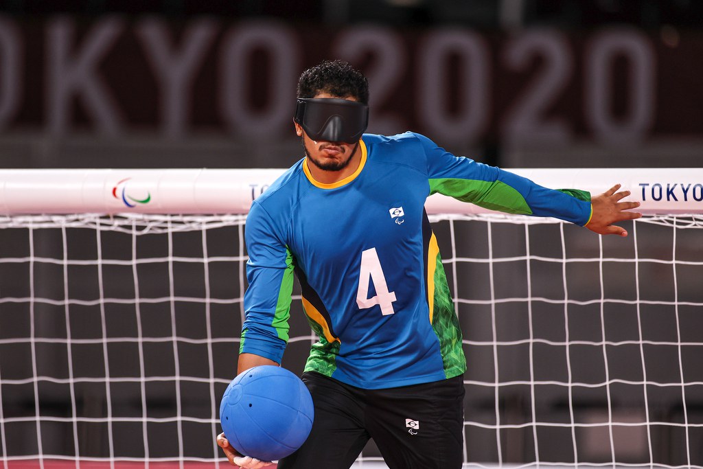 #Acessibilidade: Leomon segura a bola com a mão direita enquanto inicia o giro antes do arremesso. Ele veste camisa de manga longa azul com o número 4 escrito em branco. Ao fundo, a logo de Tokyo 2020. 